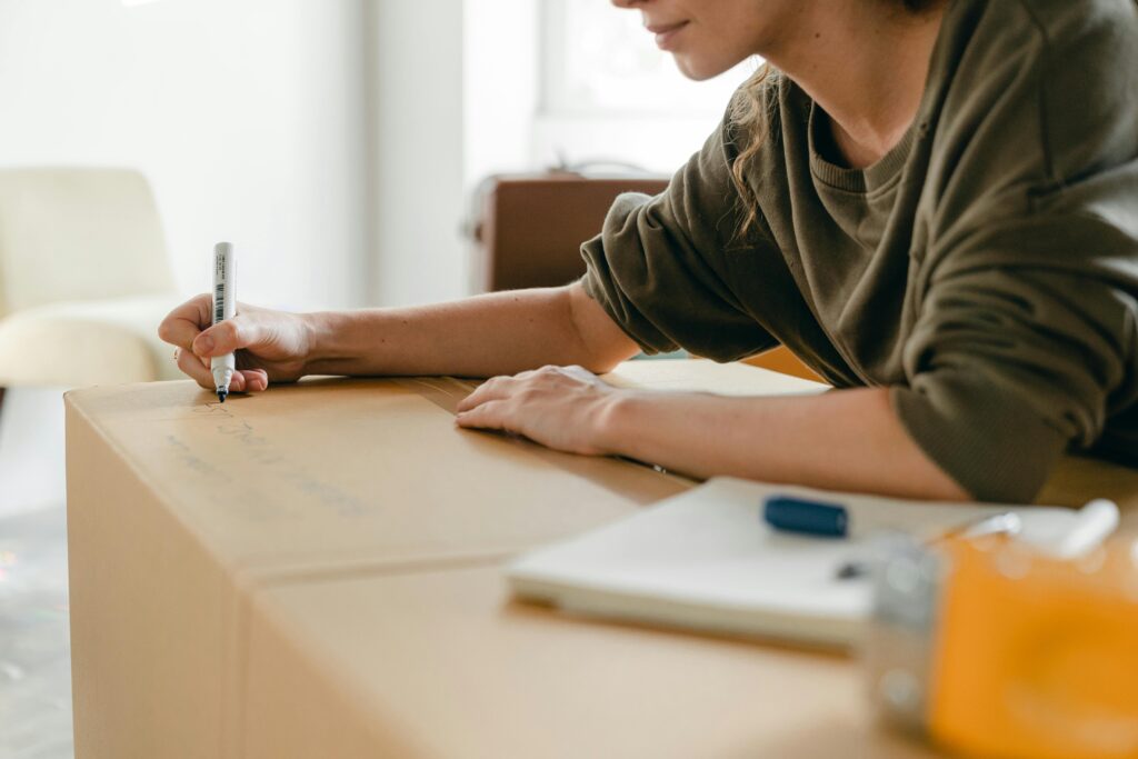 pexels-photo-4246188-4246188 Side view of crop anonymous female in casual wear writing with marker on big cardboard box while sitting near notebook in light flat