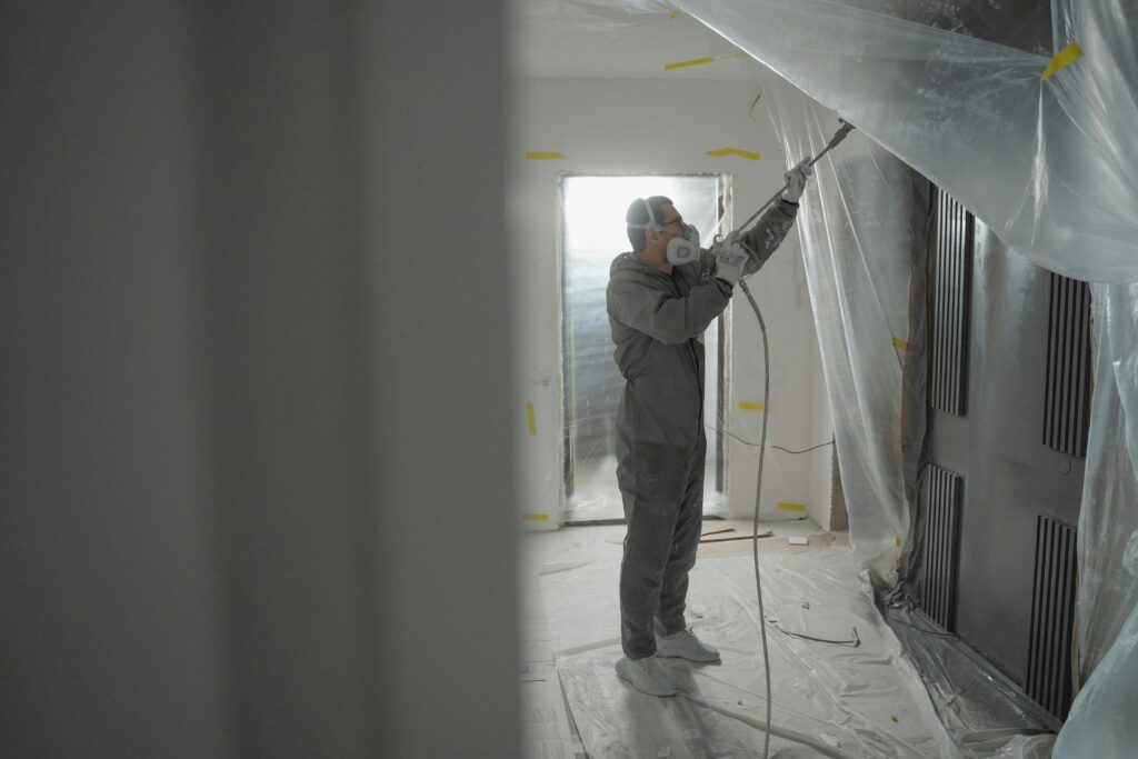 pexels-photo-6474199-6474199 Man in protective gear spray painting a house's interior during renovation work.